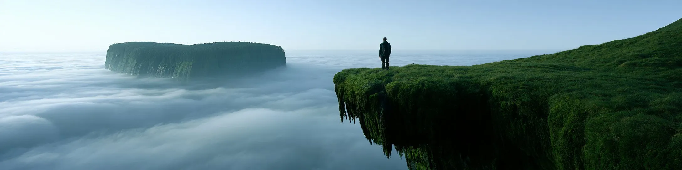 A lone hiker on the mossy edge of a plateau above a sea of cloud, another plateau floating in the distance.