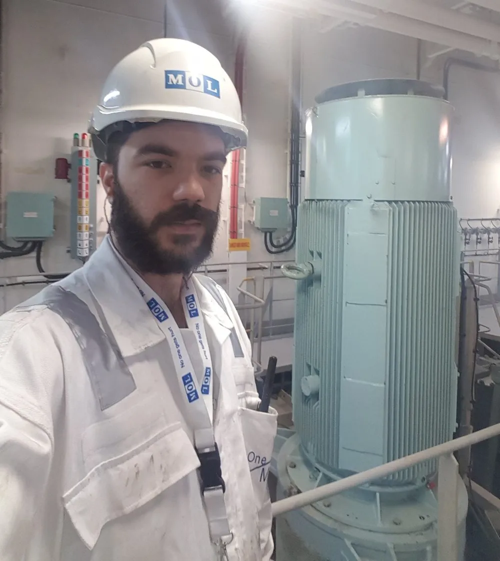 Luka in white overalls and a MOL hard hat, standing beside a large motor in a ship's engine room